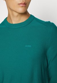 Jeune homme portant un pull ras du cou bleu sarcelle avec un col côtelé et un petit logo "BOSS" brodé sur la poitrine, regardant sur le côté.