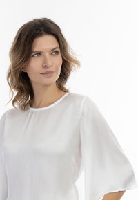 Fehér áttetsző blúz kerek nyakkivágással és laza, háromnegyedes ujjakkal. Az anyagnak finom, textúrált mintázata van.