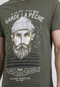 T-shirt en coton vert olive avec un graphique d'un marin barbu, un design monochrome à rayures et du texte en blanc et noir.