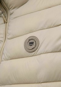 Beige Pufferjacke mit gepolstertem Material und horizontalen Nähten. Sie verfügt über einen runden Stoff-Logoaufnäher mit dem Markennamen und dem Entwurfsjahr.