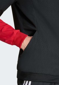 Veste noire à finition texturée avec des manches en tissu rouge et une poche latérale. Poignets élastiques et design ajusté sont des détails remarquables.