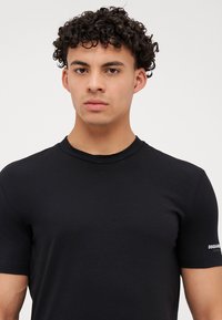 Joven con el cabello rizado que lleva una camiseta negra de manga corta lisa con un pequeño logo blanco en la manga izquierda.