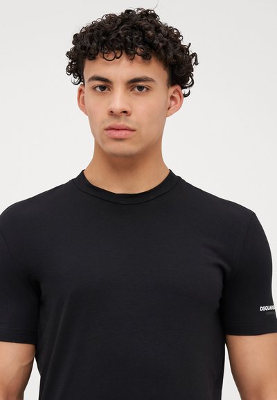 Junger Mann mit lockigem Haar, der ein schlichtes schwarzes T-Shirt mit kurzen Ärmeln trägt, das ein kleines weißes Logo auf dem linken Ärmel hat.