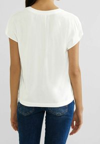 White short-sleeve top with a relaxed fit, featuring a rounded neckline and pleat detail at the back, paired with blue jeans.