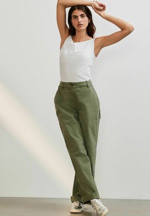Débardeur blanc sans manches avec détail drapé, associé à un pantalon cargo vert olive avec poches latérales et fermeture à bouton. Baskets argentées.