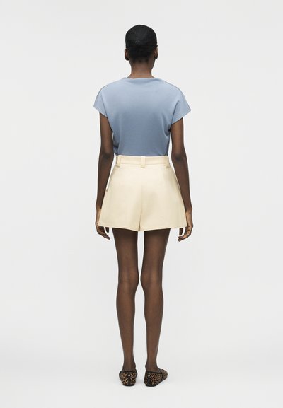 Γυναίκα που στέκεται με το πρόσωπο προς τα πίσω, φορώντας ένα ανοιχτό μπλε κοντομάνικο μπλουζάκι, κρεμ σορτς με ψηλή μέση και φλατ παπούτσια με λεοπάρ μοτίβο.