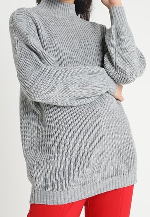 Femme portant un pull à col roulé en grosse maille gris clair à manches longues et un pantalon rouge vif, posant avec un bras plié.