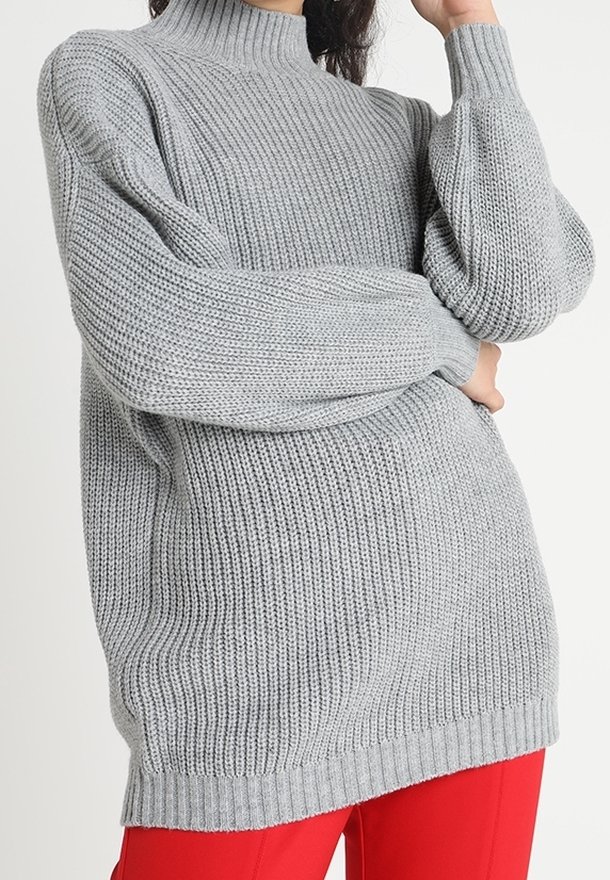 Femme portant un pull à col roulé en grosse maille gris clair à manches longues et un pantalon rouge vif, posant avec un bras plié.