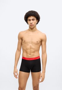 Μαύρα βαμβακερά μποξεράκια με κόκκινη μέση που φέρει το λογότυπο «TOMMY HILFIGER», κατασκευασμένα από απαλό, ελαστικό ύφασμα. Το μοντέλο στέκεται με τα χέρια στα πλάγια.