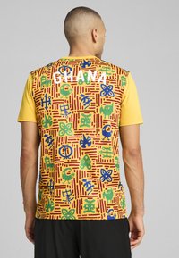 Kortærmet trøje i gul med et mønstret ryg i røde linjer og multifarvede symboler. "GHANA" er fremtrædende vist i hvid.
