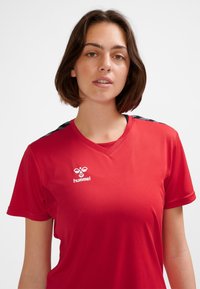 Maglietta atletica rossa con scollo a V. Presenta maniche corte e dettagli neri sulle spalle. Logo Hummel bianco sul petto.