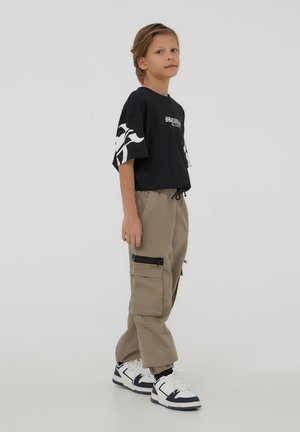 T-shirt nera con maniche grafiche bianche, abbinata a pantaloni cargo beige con tasche laterali con zip. Sneakers bianche e blu completano l'outfit.