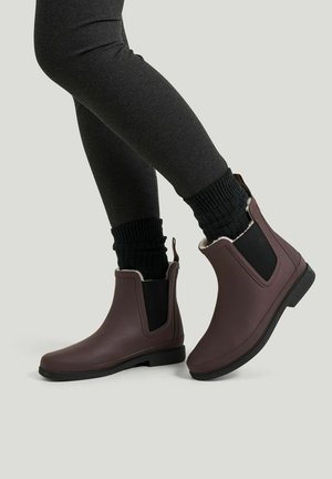 Person iført mørke leggings, sorte strikkede sokker og brune ankelregnstøvler med elastiske sidelommer og pelsfor, stående på en lys baggrund.