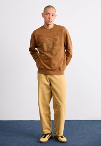 Sudadera marrón con un estampado gráfico en beige, combinada con pantalones amarillos claros y zapatillas negras y amarillas. Diseño minimalista.