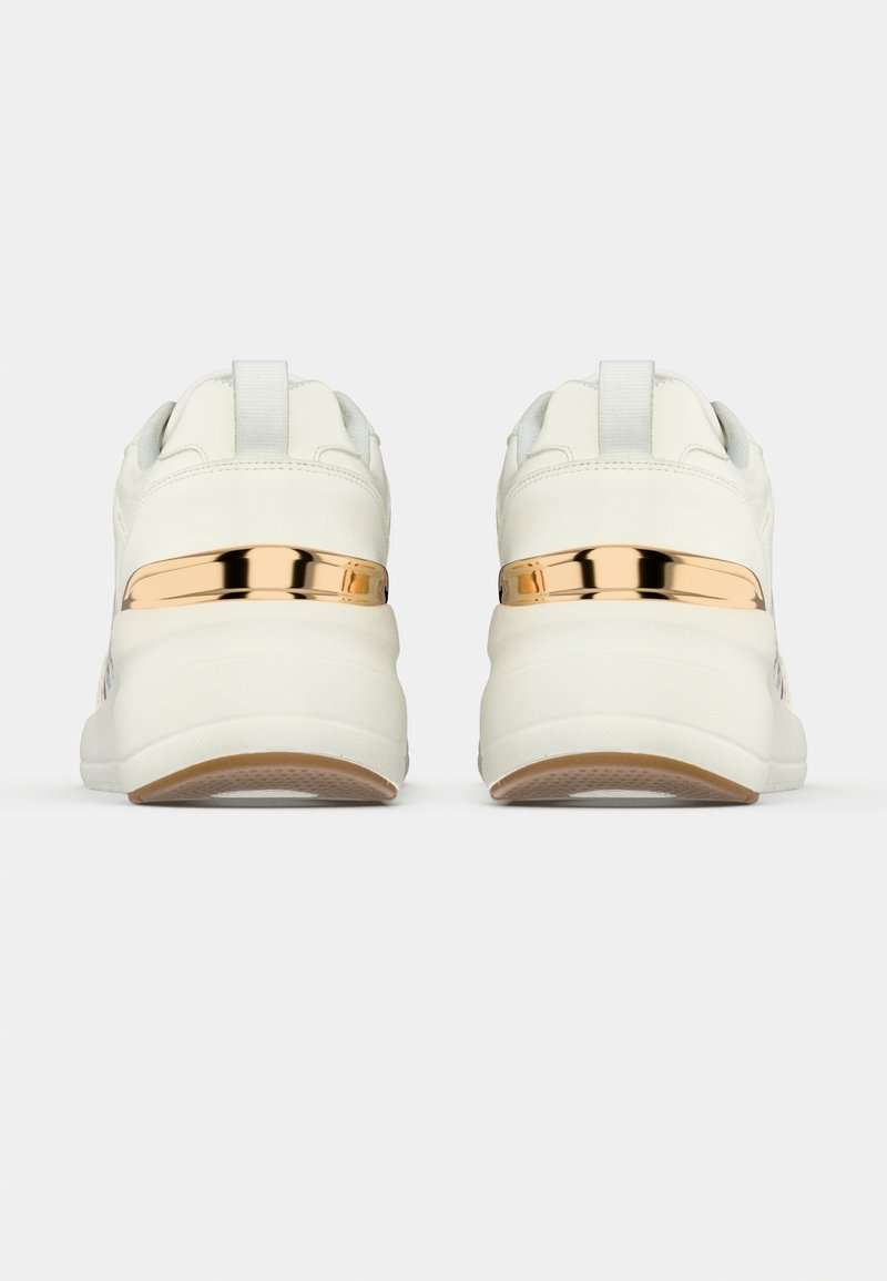 Paire de baskets blanches vues de dos avec un détail de bande métallique dorée au-dessus de semelles blanches épaisses sur un fond uni.