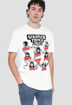 Young man wearing white Stranger Things Tales from 85 T-shirt featuring black and red illustrated character portraits and names.