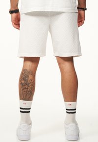 Weiße strukturierte Shorts mit einem Diamantmuster, getragen mit weißen Sneakers und Socken mit schwarzen Streifen. Ein Bein-Tattoo ist teilweise sichtbar.