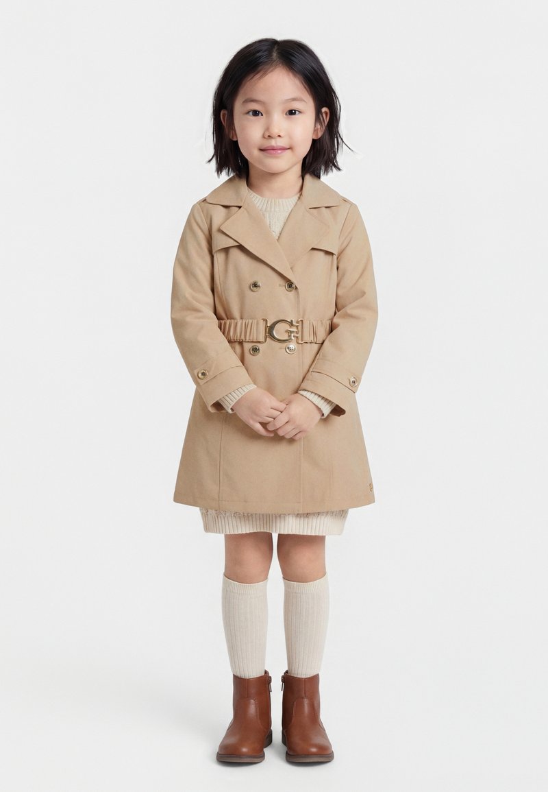 Jeune fille debout face à l'avant, portant un manteau beige ceinturé, une robe crème, des chaussettes mi-mollet assorties et des bottines marron, sur fond blanc.