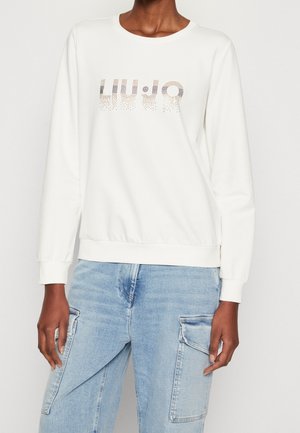 Person wearing a white long-sleeve sweatshirt with "LIU·JO" text in small metallic dots and light blue cargo jeans.