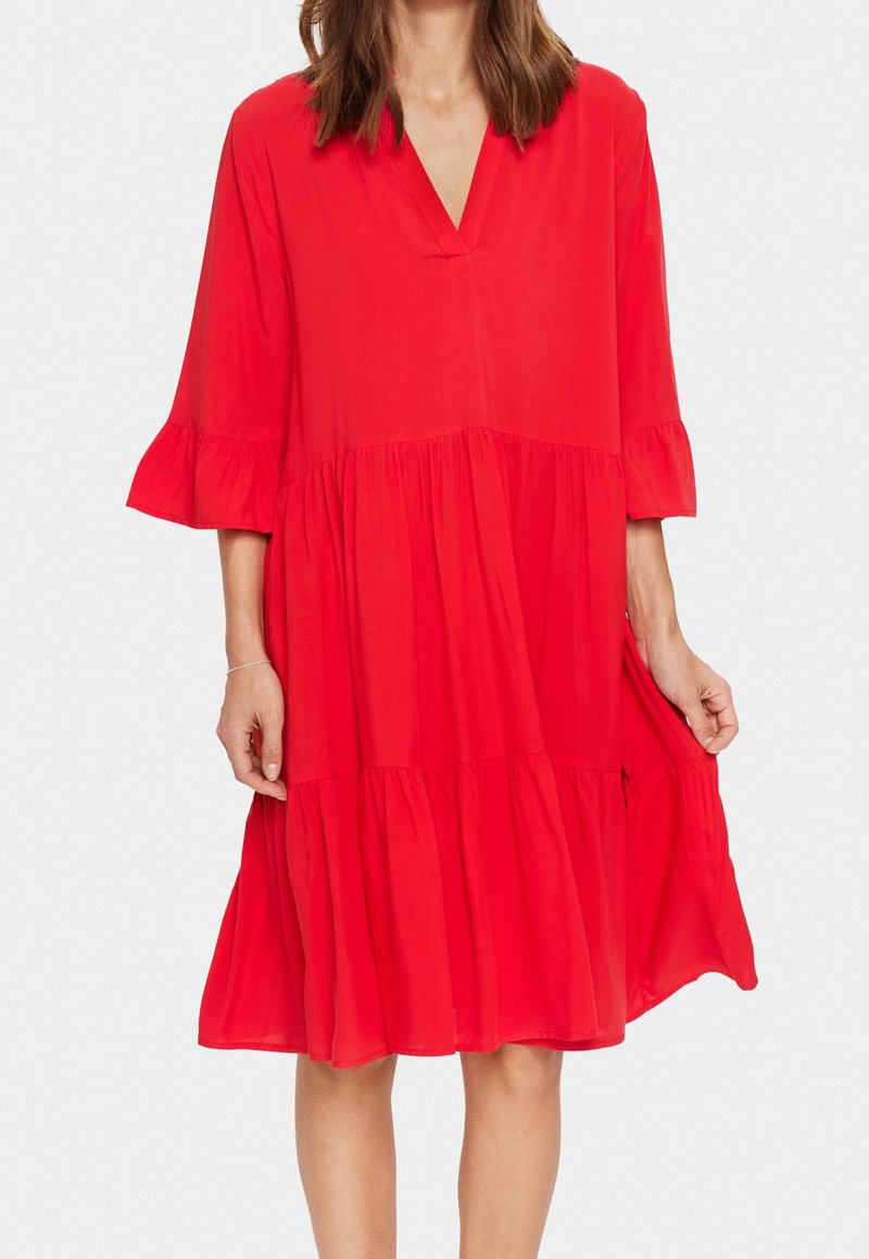 Femme portant une robe rouge ample, longueur genou, avec des manches trois-quarts à volants et un décolleté en V, tenant la robe légèrement sur le côté.