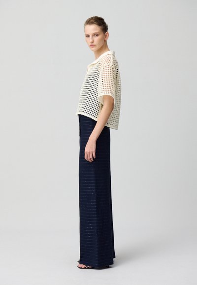 Top en crochet crème avec une coupe crop, associé à une jupe bleu marine ajustée avec des lignes subtiles. Des sandales complètent le look.