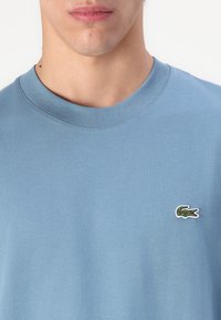 Gros plan sur le cou et le haut de la poitrine d'une personne portant un t-shirt bleu clair à col rond avec un petit logo de crocodile vert sur le côté droit.