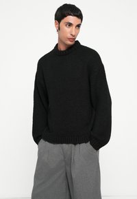 Pull en maille noir avec un col haut à côtes, des épaules tombantes et une coupe décontractée. Porté avec un pantalon gris à jambes larges.