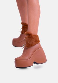 Stivali alla caviglia con zeppa in plataforma marroni, con polsini in pelliccia sintetica, punta rotonda e tacchi spessi. Finitura in pelle liscia, dettagli con lacci e accenti testurizzati.