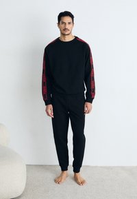 Svart hettegenser og joggebukser med rødt HUGO-logo på ermene, ribbede mansjetter og myk tekstur. Modell står barbent mot en lys bakgrunn.