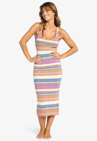 Frau steht barfuß mit den Händen in der Hüfte, trägt ein ärmelloses, geripptes, gestreiftes Kleid in Beige, Blau, Pink und Weiß vor einem weißen Hintergrund.