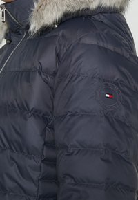 Marineblaue Steppjacke mit Kapuze aus Kunstfell. Verfügt über horizontale Steppnähte und einen runden Logopatch am Ärmel in Rot, Weiß und Blau.