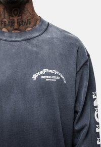 Felpa a maniche lunghe grigio scuro con una texture morbida; presenta del testo stampato bianco sul davanti e sulla manica destra; colletto a costine.