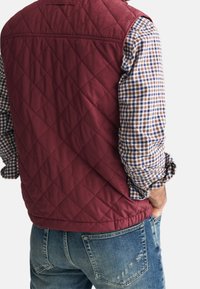 Man in een kastanjebruine gewatteerde vest over een blauw, bruin en wit geblokt shirt met opgerolde mouwen en blauwe versleten spijkerbroek, met zijn handen in zijn zakken.
