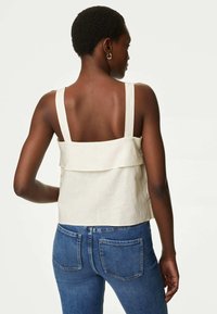 Světle béžový lněný crop top se širokými ramínky a vrstveným lemem, doplněný o tmavě modré džíny s klasickým designem zadní kapsy.