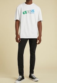 T-shirt bianco in cotone con logo "GUESS ORIGINALS" multicolore, abbinato a jeans neri in denim e sneaker alte nere.