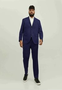Homem com barba e tranças, vestindo um fato azul-marinho, camisa branca e sapatilhas pretas, em pé contra um fundo claro e liso.