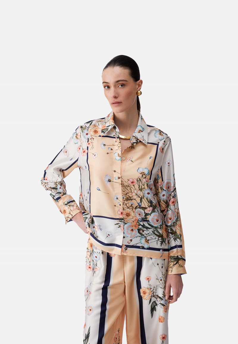 Femme aux cheveux foncés portant un chemisier en soie à motif floral et un pantalon assorti, debout avec une main sur la hanche devant un fond uni.