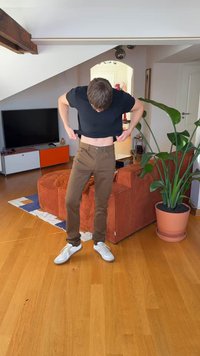 Brune, slimfit bukser med forlommer, kombineret med en sort T-shirt og hvide sneakers, inde i et rum med trægulv og en grøn plante.