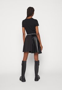 Kvinna med flätat hår som bär en svart t-shirt med korta ärmar, svart plisserad kjol med läderpaneler och knähöga svarta stövlar, stående med ryggen mot kameran.