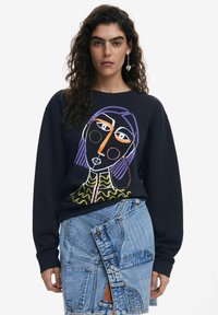 Marine sweatshirt met kleurrijke abstracte gezichtsillustratie, voorzien van lange mouwen en geribbelde manchetten. Gesteund door een denim wikkelrok met overlappende texturen.