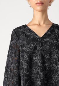 Black lace top with floral embroidery, featuring a V-neck and long sleeves, showcasing intricate patterns and textured fabric.