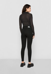 Sieviete ar cirtainiem matiem, ģērbta melnā caurredzamā garajās piedurknēs, melnās augstās šortās, kuru sānā redzama CK birka, un melnās loafers.