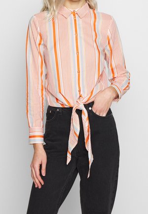 Femme portant une chemise à rayures orange, blanche et bleue nouée à la taille avec un jean noir taille haute, main dans la poche.