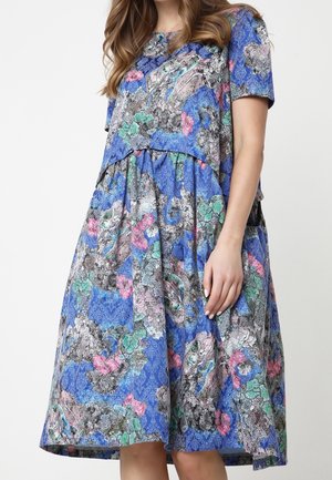 Femme portant une robe mi-longue à manches courtes avec une base bleue et un motif floral abstrait multicolore, debout devant un fond blanc.