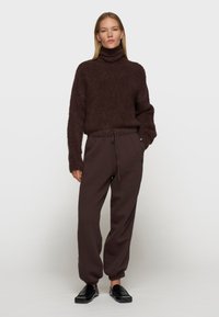 Brązowy, puszysty sweter z golfem w połączeniu z ciemnoskórą, luźno dopasowaną dresową spodnią oraz czarnymi butami wsuwanymi. Minimalistyczny design, miękka faktura.