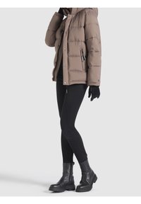 Chaqueta de plumas en marrón claro con secciones acolchadas, capucha y bolsillos laterales con cremallera, llevada sobre leggings negros y botas negras gruesas.