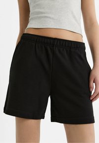 Osoba oblečená v černých elastických šortkách a šedém zkráceném topu, stojící proti čistému bílému pozadí.