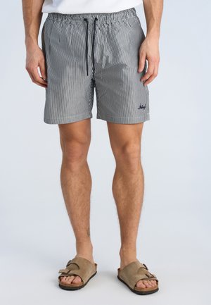 Mand iført sort-hvide smalle stribede shorts med snøre og beige sandaler på ensfarvet baggrund.