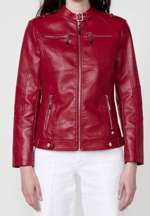 Femme portant une veste en cuir rouge cintrée avec fermeture éclair à l’avant et poches zippées, associée à un pantalon blanc.