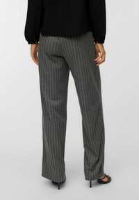 Personne portant un pantalon large à rayures grises, un haut à manches longues noir et des escarpins noirs à bout ouvert, se tenant devant un fond uni.